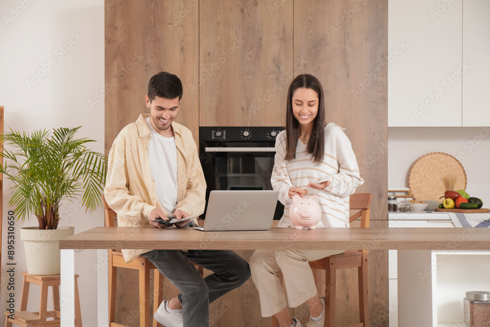 Wall mural young couple putting money into piggy bank at table in kitchen