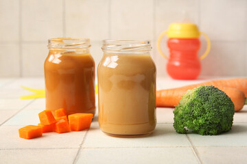 Glass jars of healthy baby food and vegetables on light tile background