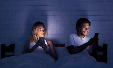 Cheating Concept. Unfaithful Husband Chatting On Cellphone Lying With Wife In Bed At Night. Selective Focus, Low Light