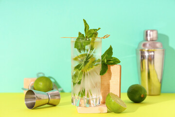 Glass of fresh mojito with jigger on yellow table against turquoise background