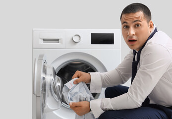 Obraz premium Shocked young businessman holding shirt with lips mark near washing machine on grey background