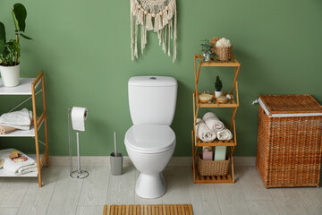 Toilet bowl, shelving unit, paper holder and laundry basket near green wall in restroom
