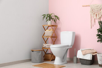 Toilet bowl, shelving unit, bin and houseplant near pink wall in restroom
