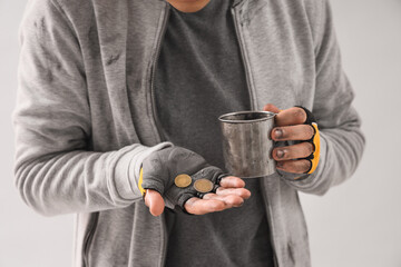 Young homeless man with cup and coins on light background, closeup