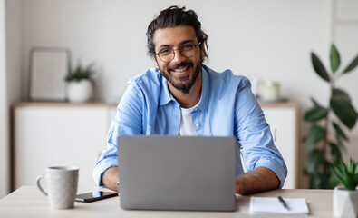 Successful Freelancer. Happy Millennial Indian Man Sitting At Desk With Laptop, Smiling And Posing At Camera While Working On Computer At Home Office, Wearing Eyeglasses And Braces, Free Space