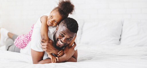 A happy black father lies on a bed with his daughter on top of him, both smiling brightly at the...