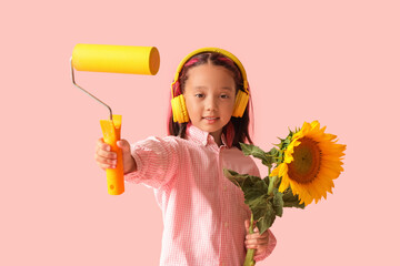 Cute little Asian girl in headphones with beautiful sunflower and paint roller on pink background