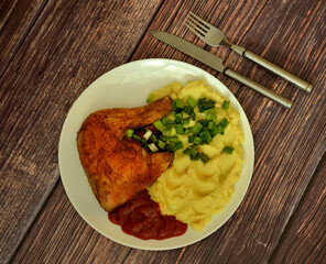 A plate of mashed potatoes, baked chicken leg and tomato sauce on a wooden table, with cutlery nearby.