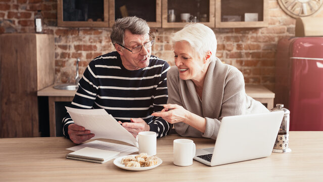 An older couple sits at a kitchen table, reviewing documents while enjoying a cup of coffee. The man is holding a paper in his hands while the woman points to something on the paper.