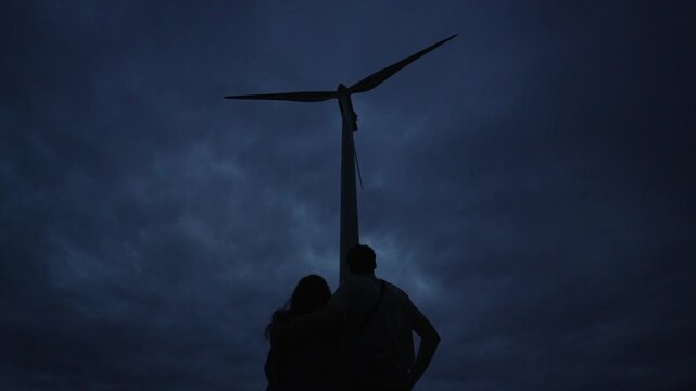 Silhouette of two people standing in front of a windmill in the dark, looking up in the dark clouds, static shot with copy space, windmill not moving