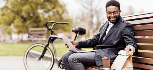 Obraz premium A professional black man uses his laptop on a park bench with a bicycle nearby. The atmosphere is serene and efficient, capturing the essence of outdoor productivity.
