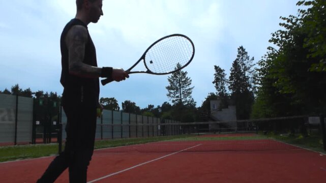A man with tattoos flipping a tennis racket in a tennis court during the summer and day time in 4K. Footage features beautiful weather, sunshine and cloudy sky with tennis fields in the background.