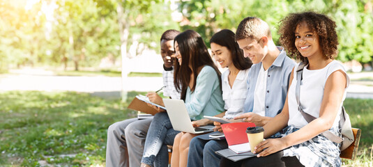 Group of students studying together outdoors, preparing for lectures and classes while sitting on bench in campus, black girl smiling at camera