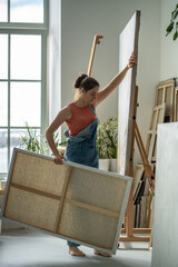 Woman artist carrying large canvas in hands in home art studio. Creative female artist works on...