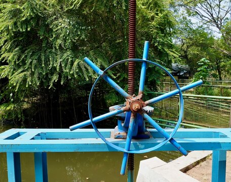 Metal iron blue colored water way or gate door wheel spinner isolated on horizontal green pond water and botanical plants background. - Powered by Adobe