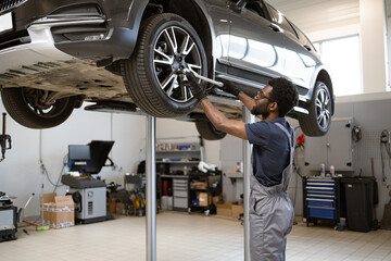 Mechanic wearing overalls working on car lifted for maintenance in modern automotive repair shop. Maintenance tools and equipment visible in clean and organized workspace.