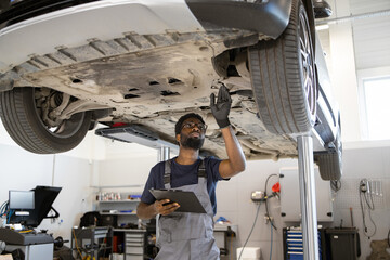 Mechanic conducts vehicle inspection while car is elevated on hydraulic lift in modern auto repair shop environment. Professional examines undercarriage, ensuring maintenance and safety standards.