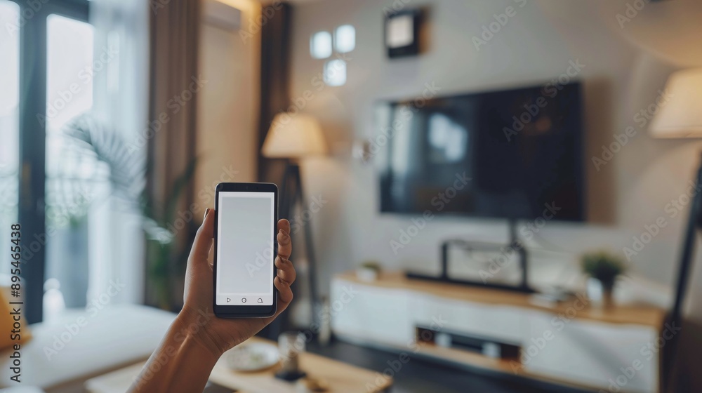 Poster POV image showing a hand holding a mobile phone with a blank white screen near a smart home device like a thermostat Stock Photo with copy space
