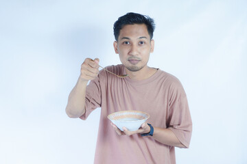 A portrait of an Indonesian Asian man wearing a pink tshirt, seen happily posing and enjoying his meal with a spoon and bowl, isolated on a green background.