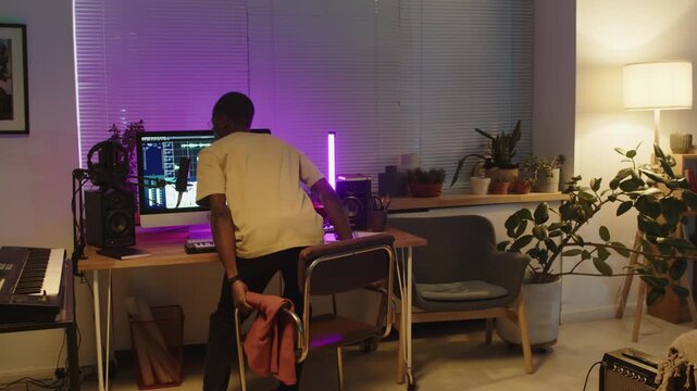 Young African American musician walking in home recording studio with trendy neon light, sitting at desk and putting headphones on