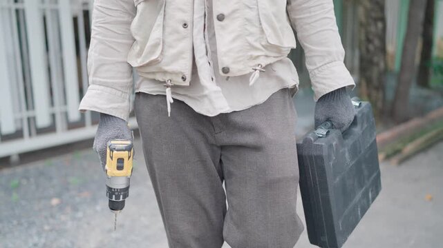 Construction tools, young asian carpenter in grey gloves walking with drill and toolbox to do woodwork or repair, drilling with drill, home building, craftsman, carpenter, craft, diy, construction,