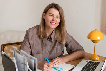 Young woman working or studying at laptop, writing in notebook. Looking at camera and smiling.