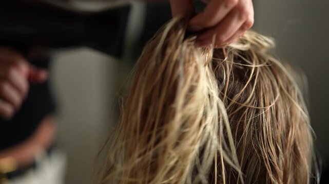 Closeup of female hairdresser accurate drying wet long blonde hair of young woman customer with hairdryer in beauty studio. Lady client getting stylish hairdo done at salon. Shooting in slow motion.