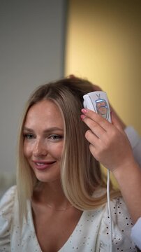 Cheerful blonde female visiting trichologist in dermatological clinic. Closeup hands of unrecognizable dermatologist making diagnosis of structure and condition of hair with special device trichoscope