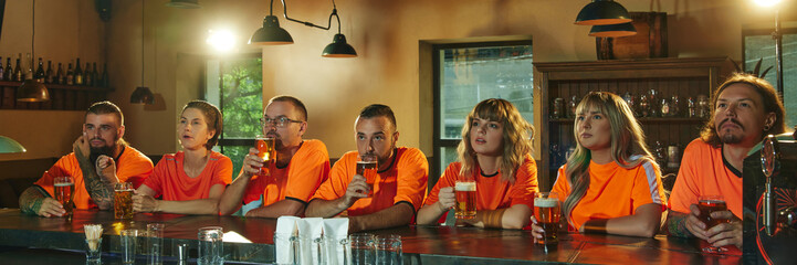 Banner. young people, women and men, in bright orange shirts sits and intensely watching TV and drinks beer at bar. Concept of friendship, soccer fan, championship, support, celebration. Ad