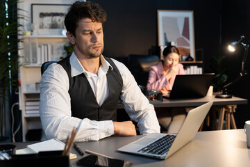 Sitting man with serious face thinking creative business project planning at night lighting time at modern office with blurry secretary checking paper report with folder background concept. Postulate.