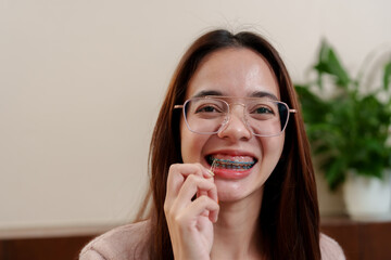 Asian teenage girl with braces, smiling and flossing teeth with braces. Wearing glasses, indoors...
