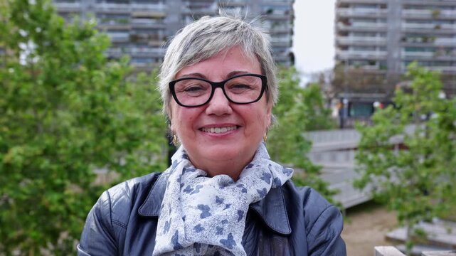 Smiling portrait of senior woman looking at camera outdoor. Head shot of 60 year old woman over urban background.