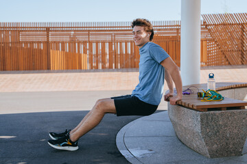 Young man doing tricep dips on a bench, smiling, wearing a blue shirt and black shorts. Water bottle and resistance bands on bench. Outdoor fitness, sunny day, healthy lifestyle