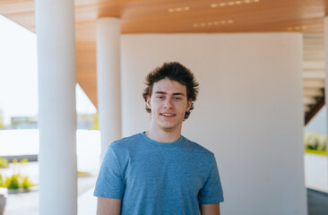 Portrait of a young man in a blue shirt standing under a modern wooden structure, smiling at the camera. Casual, confident, outdoor