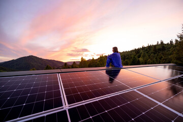 Rooftop with solar panels on house in mountains, woman sitting alone enjoying sunset. Energy...