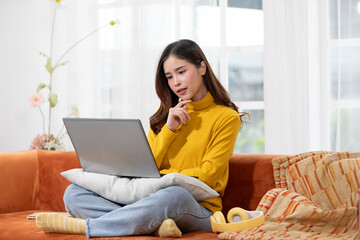 Asian young woman in a yellow sweater sits on a couch, using her laptop with a thoughtful expression. She is comfortably positioned with a pillow on her lap