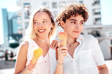 Two young beautiful smiling hipster female in trendy summer clothes. Carefree women posing on street background. Positive models eating tasty ice cream in waffles cone in sunny day, cheerful and happy