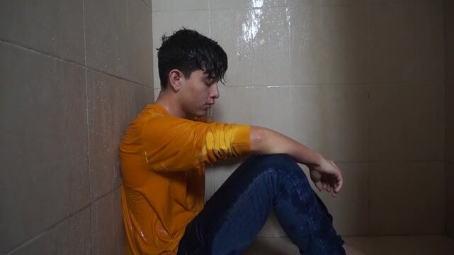 A young man sits hunched in the corner of the shower, fully clothed. The water streams over him, mixing with his tears as he stares blankly at the wall. His clothes cling to his shivering frame.