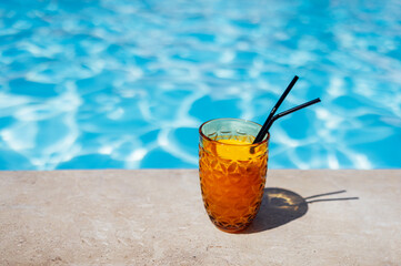 Delicious chilled fresh orange juice with straws on the side of the pool. Tasty citrus cocktail on the background of clear blue water.