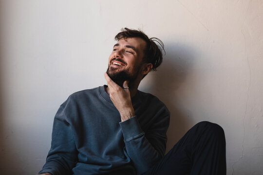 Sexy young man wearing casual grey pullover, silver ring, black pants, touch beard, holding hand to chin and sensually posing, flirting and laughing. Happy man look at side. Teeth smile male. - Powered by Adobe