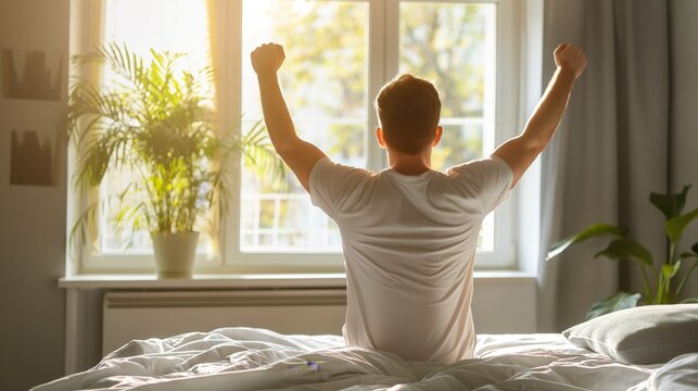 Positive excited young man enjoying good morning in cozy home bedroom, sitting on bed, feeling great after sleeping enough on comfortable mattress