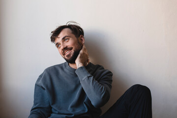 Fototapeta premium Sexy young man wearing casual grey pullover, silver ring, black pants, touch beard, holding hand to chin and sensually posing, flirting and laughing. Happy man look at side. Teeth smile male.
