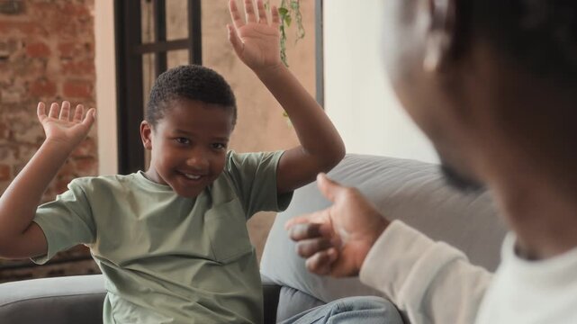 African American father and boy sitting together on couch and hanging out. Black skinned man and his son, having fun playing charades game, feeling happy, smiling and laughing.