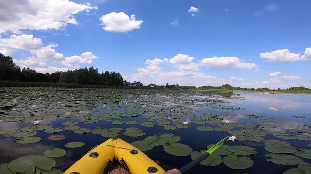 Kayaking along river through growing white lily nymph on sunny summer day. Person floating in boat on river with surface of water reflecting white clouds on calm day. Tourism. Active lifestyle. POV