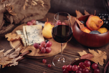 Glass of red wine served with blue cheese on dark wooden background. Autumn picnic with wine and cheese platter, fruits and dry leaves in rustic style
