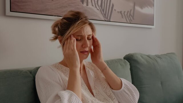 Pregnant woman sitting on sofa suffering for headache or catching a cold. Parent to be female feeling sick with hurting head. Lady in maternity massage herself.