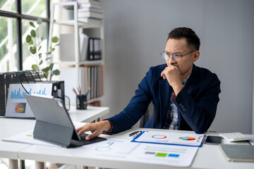 A man in a suit is working on a laptop in front of a white desk