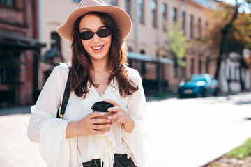 Lifestyle portrait of young stylish woman dressed in white blouse. Model walking with coffee cup in old town in sunny day. Beautiful smiling female in hat. Cheerful and happy. Takeaway coffee