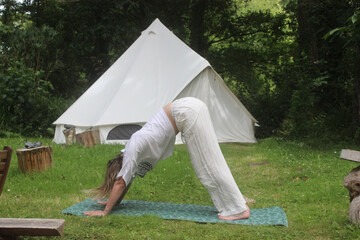 Une jeune femme fait du yoga à l'exterieur. Elle est en position du chien tête en bas . Une tente type tipi est visible à l'arrière plan © lena