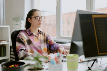 Young Caucasian female programmer dressed in spotted blouse sitting at work desk while typing on...